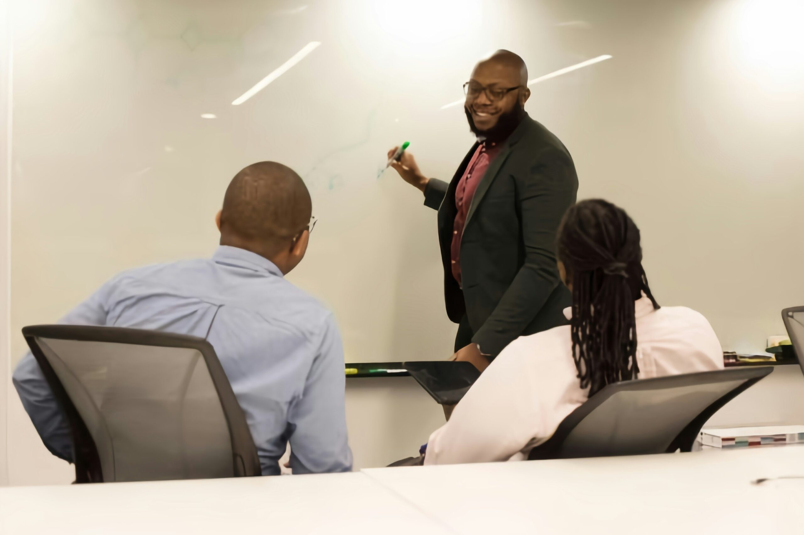 Manager Leading a Team Discussion at the Whiteboard Manager in a dark blazer writing on a whiteboard while leading a discussion with two colleagues seated at a conference table.